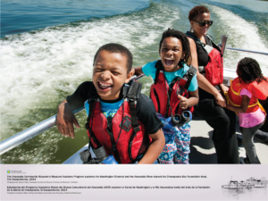 Two elementary school children wearing life jackets and binculars are shown smiling aboard the Chesapeake Bay Foundation boat while exploring the Washington Channel on a warm sunny day. An adult chaperon and another child are in the background.