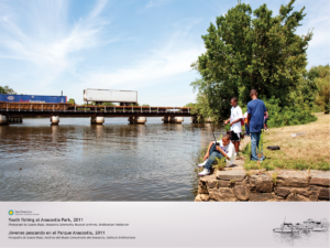 Three young men are visible sitting and standing along the stone wall bordering the Anacostia River, casting their fishing rods, on a warm sunny day. A cargo train is crossing the river in the background.