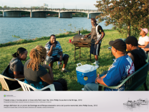 A group of seven adults are sitting in chairs, in a circle, smiling and talking with one another. One gentleman is standing and operating a grill/smoker. It appears to be nearing sunset on a warm sunny day. The Anacostia River, and a bridge, are visible in the background.