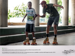 A young man and a boy are smiling as they skate together at the skating pavilion in Anacostia Park. They are wearing quad skates. It appears to be a warm and overcast day. The pillars of the pavilion are visible in the background.