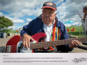 An older gentleman wearing a baseball cap, blue fleece jacket, and traditional folklorico attire is featured playing an electric guitar at the Festival del Río Anacostia.