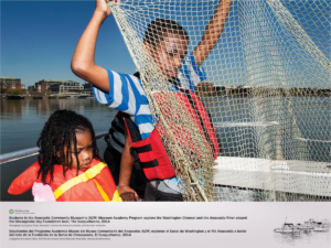 Two children are shown on a boat on the Anacostia River. A young girl wearing a life jacket sits to the left on a young boy displaying a net larger than his body. The backdrop features the waterfront in Navy Yard, Washington DC.