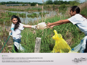 Two young women are picking up trash at Bladensburg Waterfront Park. They are standing within the wetlands, passing along a plastic container to be placed in trash bags.