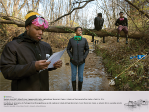 In the foreground, a young man wearing pink googles, holds a water sample and a piece of paper while standing in the creek. A young woman smiles standing behind him. In the background, two young men are seen sitting on a fallen tree, hanging over the creek. It appears to be a cooler day. All youth are wearing jackets and boots.