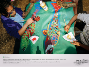 The photo shows a birds eye view of a young girl painting a fish on a picnic table outdoors. Other hands are visible, suggesting her peers are participating. Two nearly-completed fish are painted on a large piece of green paper, covering the table.