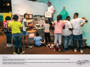 Eight students are interacting with Smithsonian Anacostia Community Museum's Reclaiming the Edge Exhibit. This section appears to be featuring the Seafarer's Yacht Club. A cutout of a sailor is holding a sign "It's Our River" as part of the exhibit.