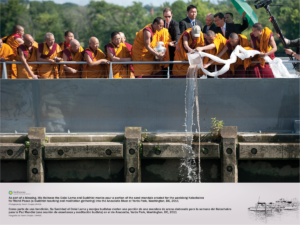 Fourteen Monks and the Dalai Lama are gathered along the Anacostia River, over a rail. The Dalai Lama poors a portion of sand mandala into the river. Five gentlemen in suits are visible in the background, with a camera visible filming the moment on the far right.