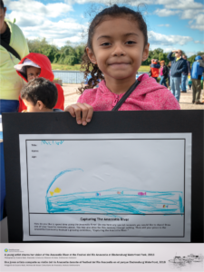 A young girl stands holding her drawing, at the Festial del Río Anacostia. Her drawing outlines her vision for the Anacostia River and includes fish swimming in water. Festival participants are visible behind her.