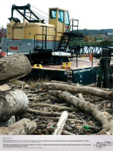 An Army Corps of Engineers clean up ship is docked along the Anacostia River on an overcast day. Wooden debris and tree trunks are scattered in the foreground.