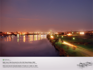 A view of the Anacostia River front the 11th Street Bridge at dusk. The Anacostia Bike Trail is shown on the righthand side, lite up with street lamps. City lights are visible on the lefthand side, exptending through the background of the image.