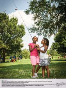 Two elementary school girls are holding a fishing rode, looking upwards at the bobber. Trees frame the girls on the left and right sides. Community members are seen enjoying the park in the background.