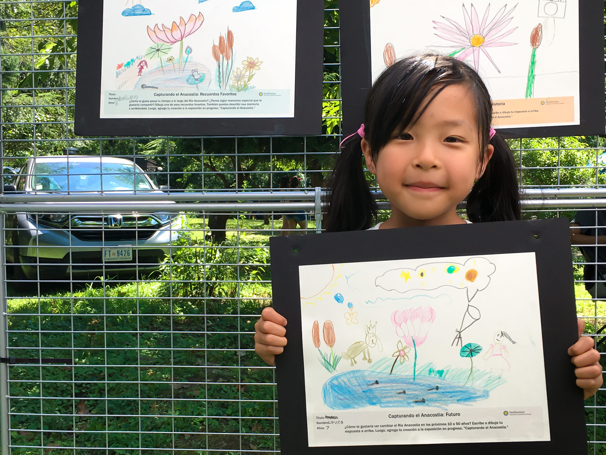 Young girl holds her drawing capturing her favorite memory of Lotus flowers in bloom while at the Kenilworth Aquatic Festival. Other drawings, displayed as part of the exhibit, are in the background.