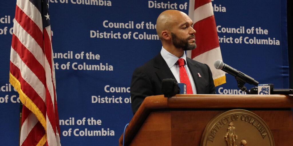 Council member Robert White stands behind a podium, next to an American Flag, as he gives his speech.