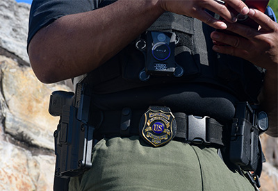 Brian Askew, in his uniform, wears a bulletproof vest with a body camera. Visible on his duty belt is a handgun, badge, magazine holder, and Taser.