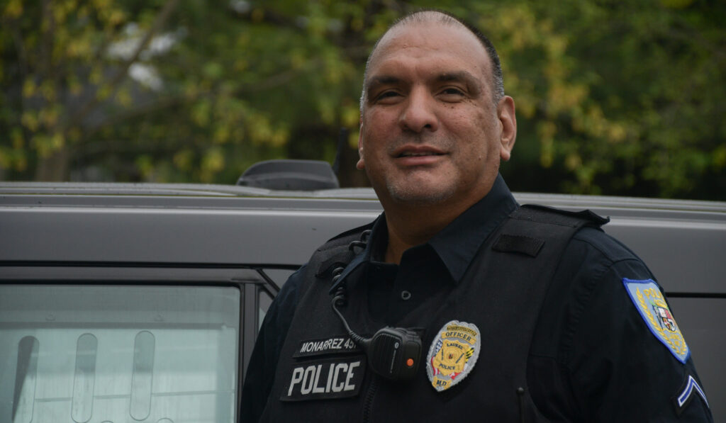 Officer Edwin Monarrez, in uniform, stands for a portrait in front of his police vehicle while on shift at the Laurel Police Department early in the morning during an overcast day.