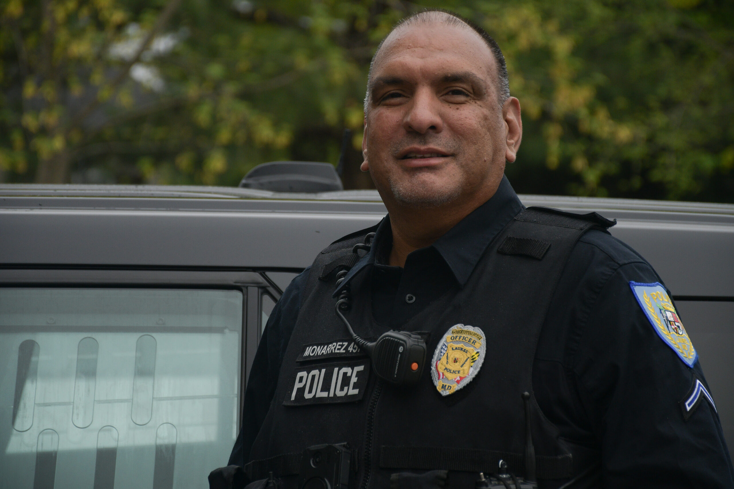 Officer Edwin Monarrez, in uniform, stands for a portrait in front of his police vehicle while on shift at the Laurel Police Department early in the morning during an overcast day.