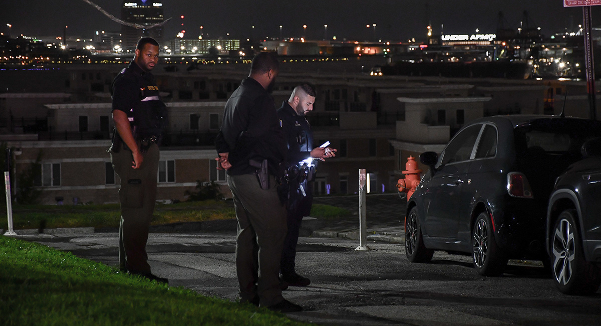 Askew and Colyns speak with a Baltimore Police officer late on a Saturday night next to Federal Hill Park. The Baltimore City skyline glistens in the background.