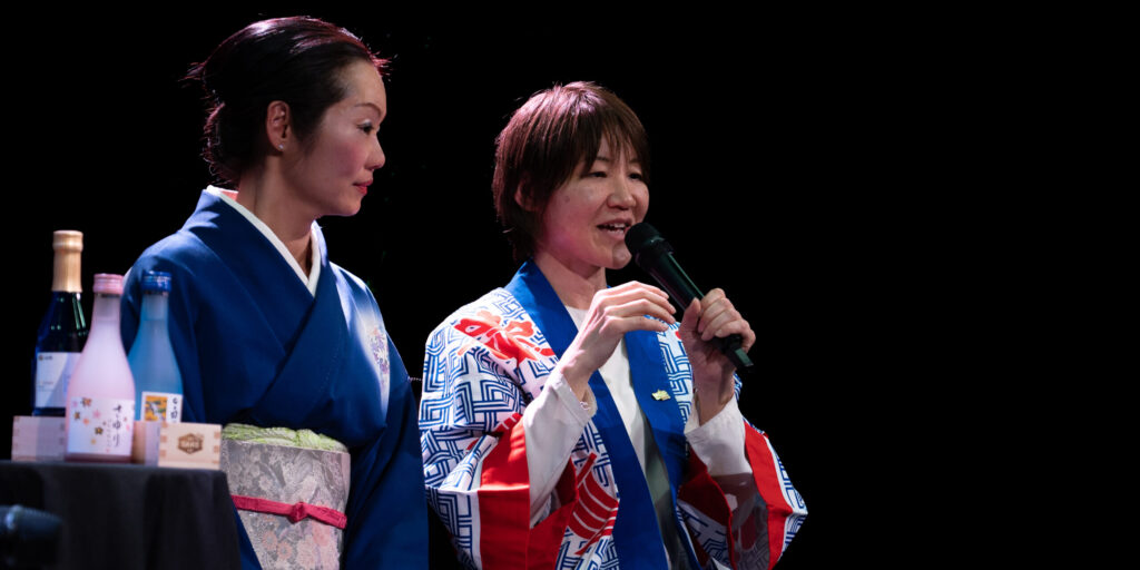 Two Japanese American Women sitting together on stage. One woman is speaking into a microphone.