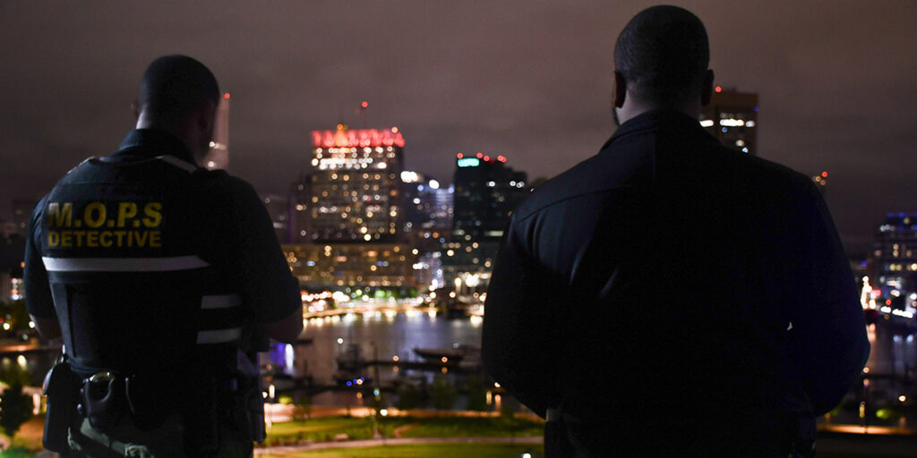 Brian Askew and Lt. Aaron Colyns, in uniform, stand side by side facing the Baltimore cityscape at night, backs to the viewer, vigilant and watchful.