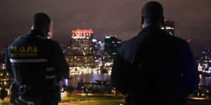 Brian Askew and Lt. Aaron Colyns, in uniform, stand side by side facing the Baltimore cityscape at night, backs to the viewer, vigilant and watchful.