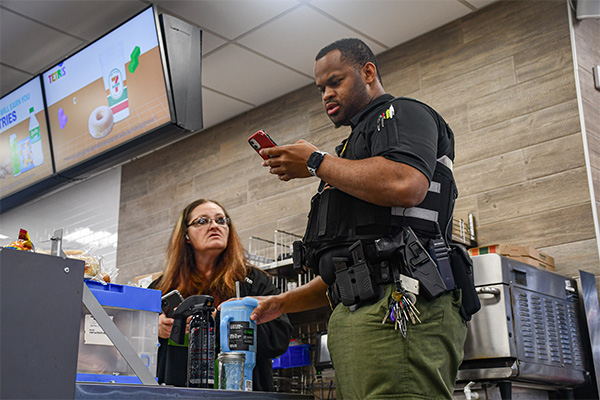 Brian Askew, in uniform, stands at the 7-11 counter looking at his cell phone while on duty. Mary Rowe is on the opposite side of the counter, conversing with Askew.