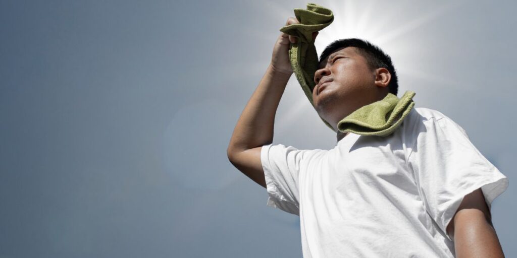 A worker looks upward, using a rag to wipe sweat off his face, with the sun blazing in the background.