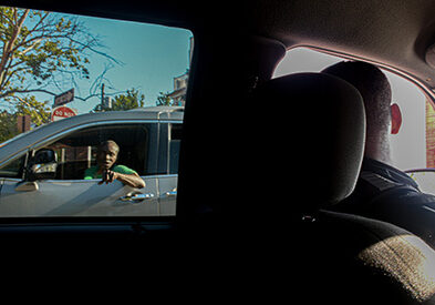 Askew is in his car, looking out the front window, engaging with an individual stopping by as he drives his car in the opposite direction.